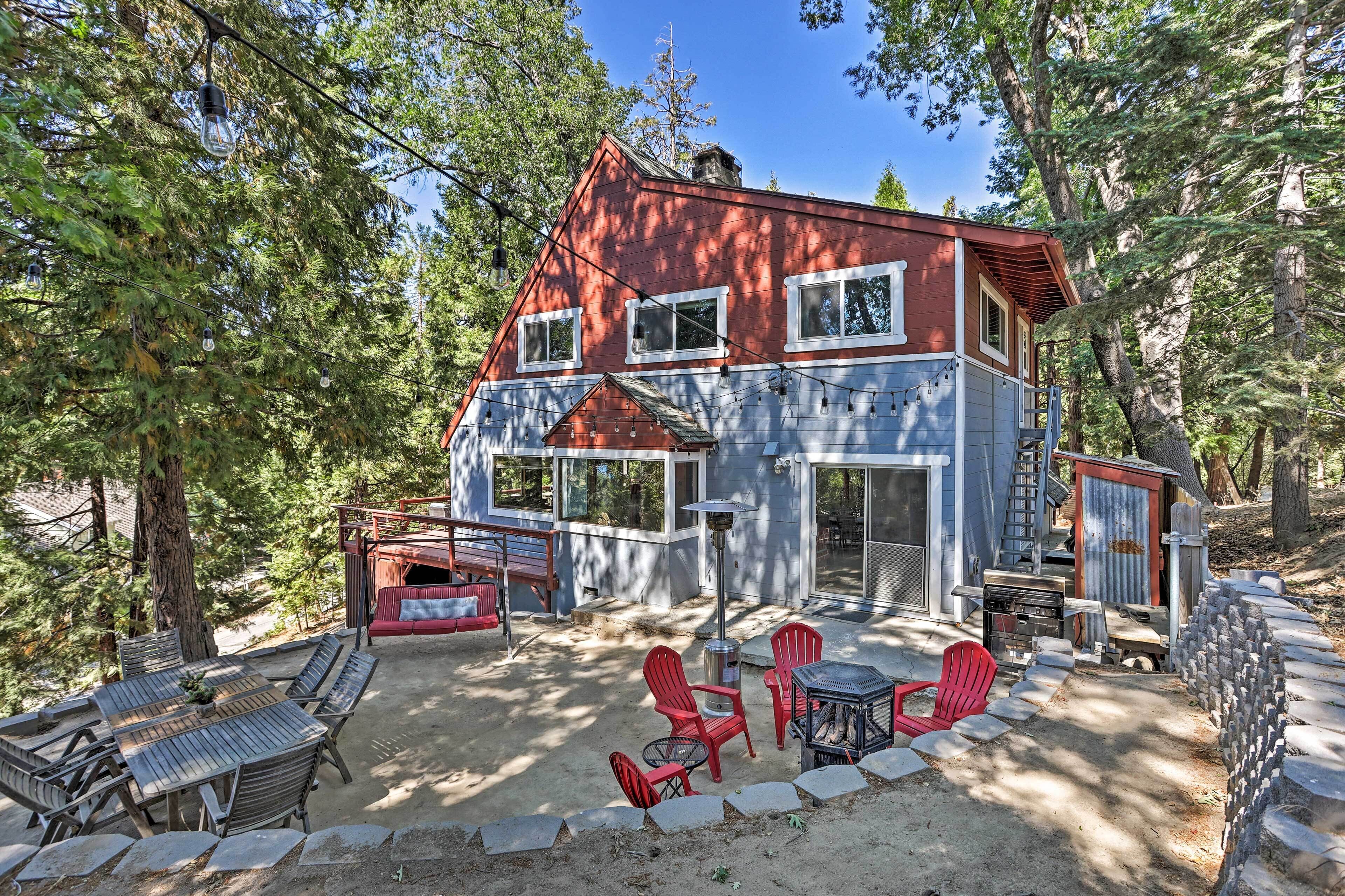 A two-story cabin with a red roof, surrounded by trees. It features a patio with outdoor furniture and a fire pit.