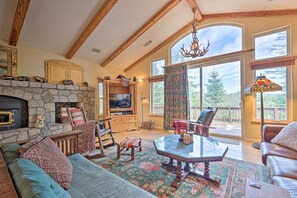 A spacious living room with a stone fireplace, wooden beams, a large window with a view of trees, and a cozy seating area. - Woodland Cabin in Lake Arrowhead: W/two Decks (Lake Arrowhead)