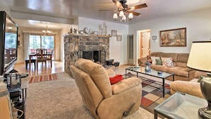 A spacious living room with a stone fireplace, a ceiling fan, and a large window overlooking a deck. - Walk to Lake: Rustic Home w/ Deck in Fawnskin (Fawnskin)