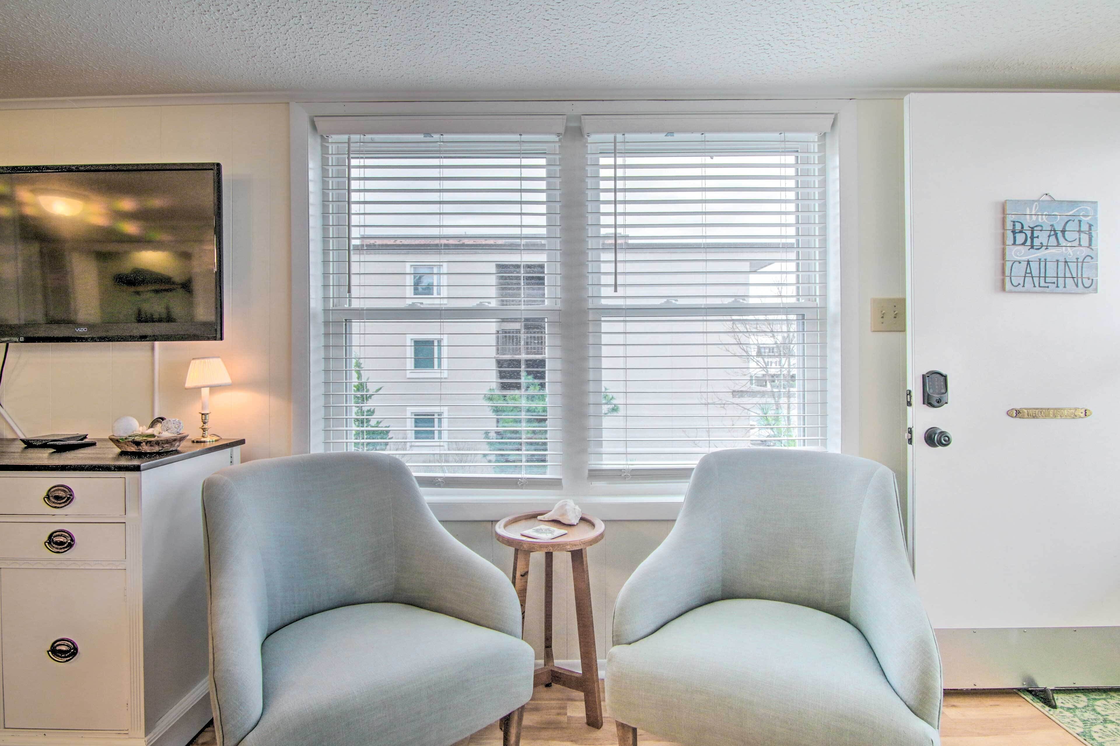 A room with two light grey armchairs, a small wooden side table, a TV on a white cabinet, and a window with blinds.
