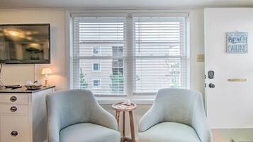 A room with two light grey armchairs, a small wooden side table, a TV on a white cabinet, and a window with blinds.