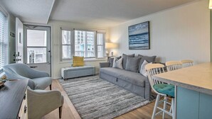 A modern living room with a grey sofa, a small ottoman, a dining area with a bar, and a large window with blinds. - Walk to Maryland Beach: Coastal Condo w/ Balcony (Ocean City)