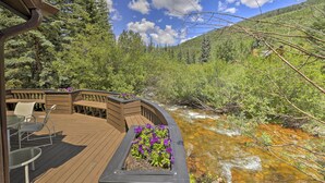 A wooden deck with outdoor seating and flower planters, overlooking a flowing stream and forested hillside. - Vail Condo w/ Mtn-view Deck - Steps to Ski Shuttle (Vail)