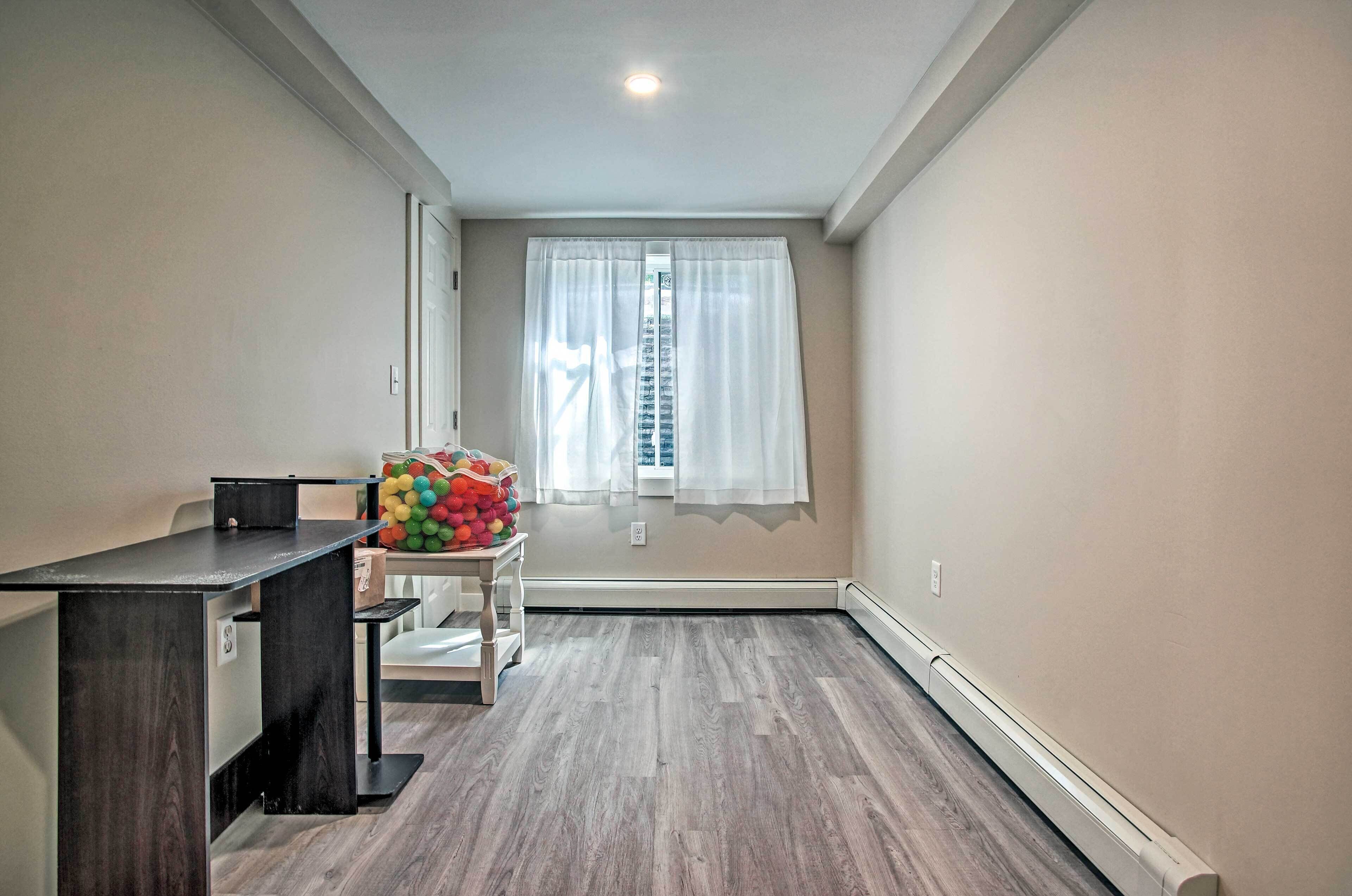 Quarto com piso de madeira, uma escrivaninha com cadeira, uma mesinha com bolas coloridas e uma janela com cortinas brancas.