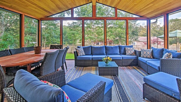 Een overdekt terras buiten met een houten plafond, grote ramen, een eettafel en blauwe gestoffeerde zitplaatsen.