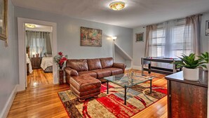 Ein Wohnzimmer mit einem braunen Ledersofa, einem Glastisch und einem roten und beigen Teppich. An der Wand hängt ein Gemälde, ein Lampenschirm und auf einem Schrank steht eine Pflanze.