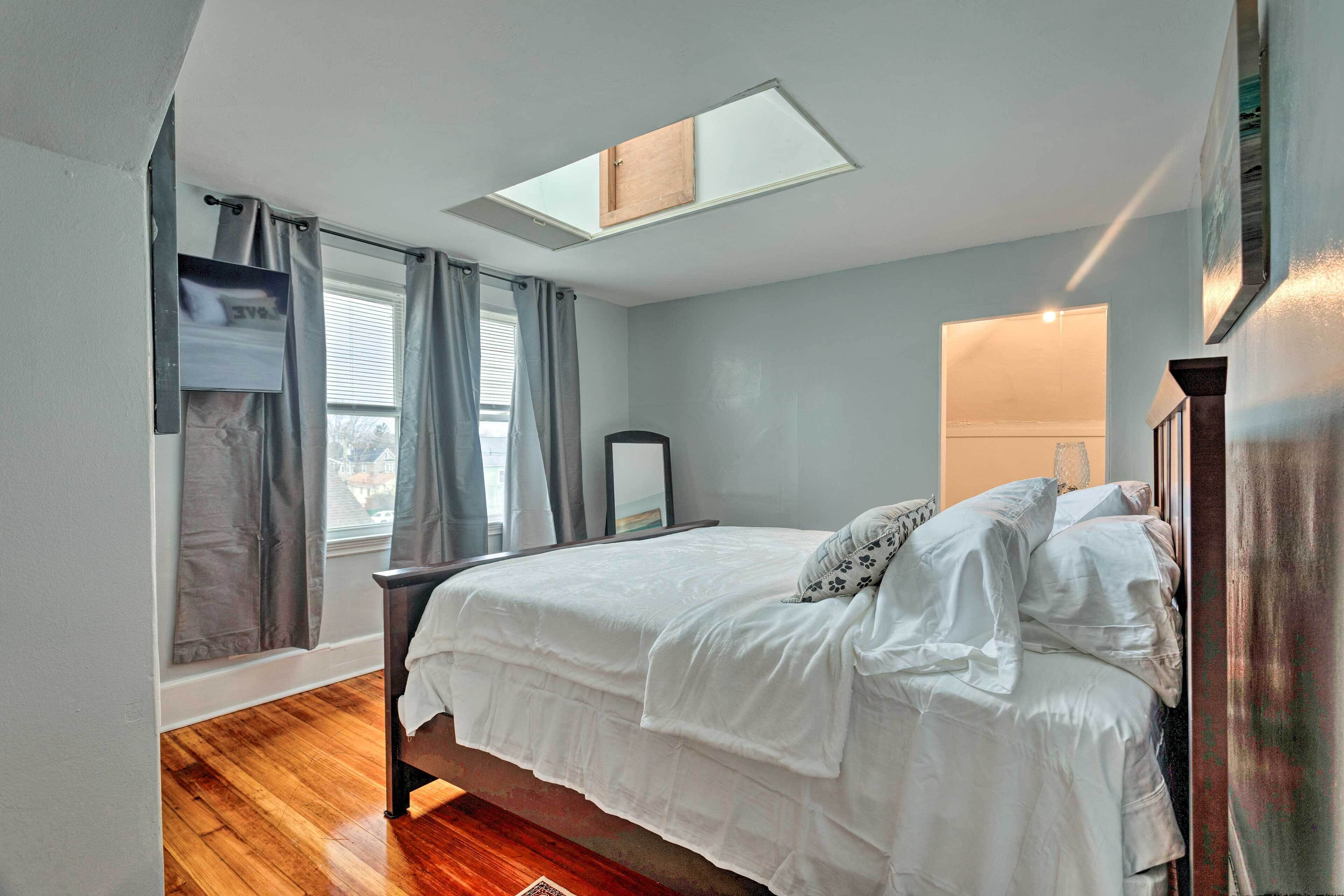 A bedroom with a bed, a skylight, a window with curtains, and a nightstand.