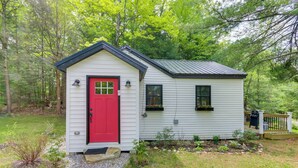 Une petite cabane à un seul étage, avec une porte rouge, un petit porche et un petit jardin.