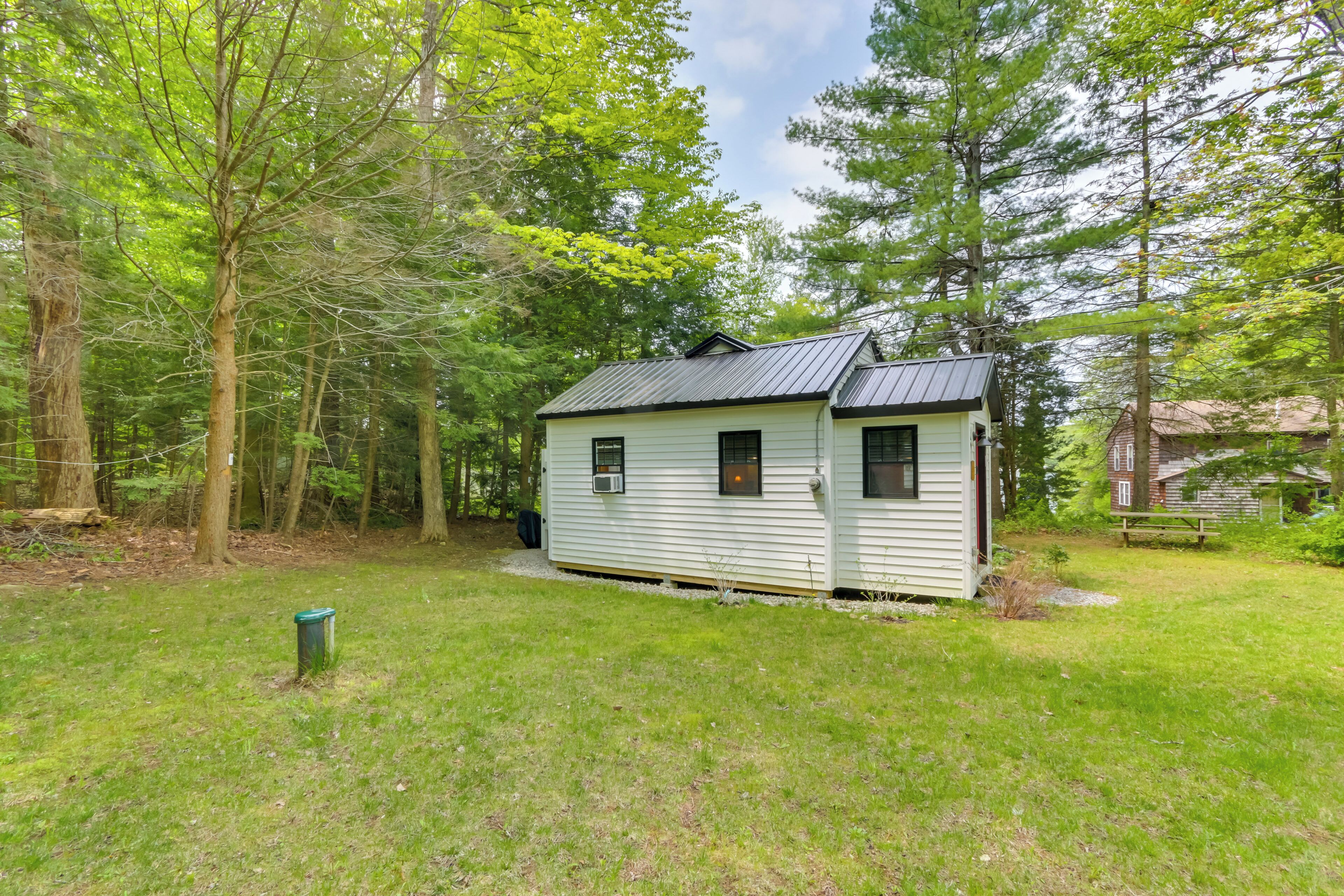 A small, single-story cabin with a metal roof, surrounded by a grassy yard and dense trees.
