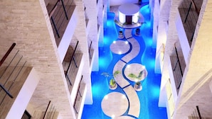 Un área de piscina interior moderna con un elemento central de agua, rodeada de zonas de estar y plantas en macetas.