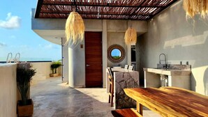 Un área de cocina exterior moderna con un banco de madera, encimera de mármol y una lámpara colgante.