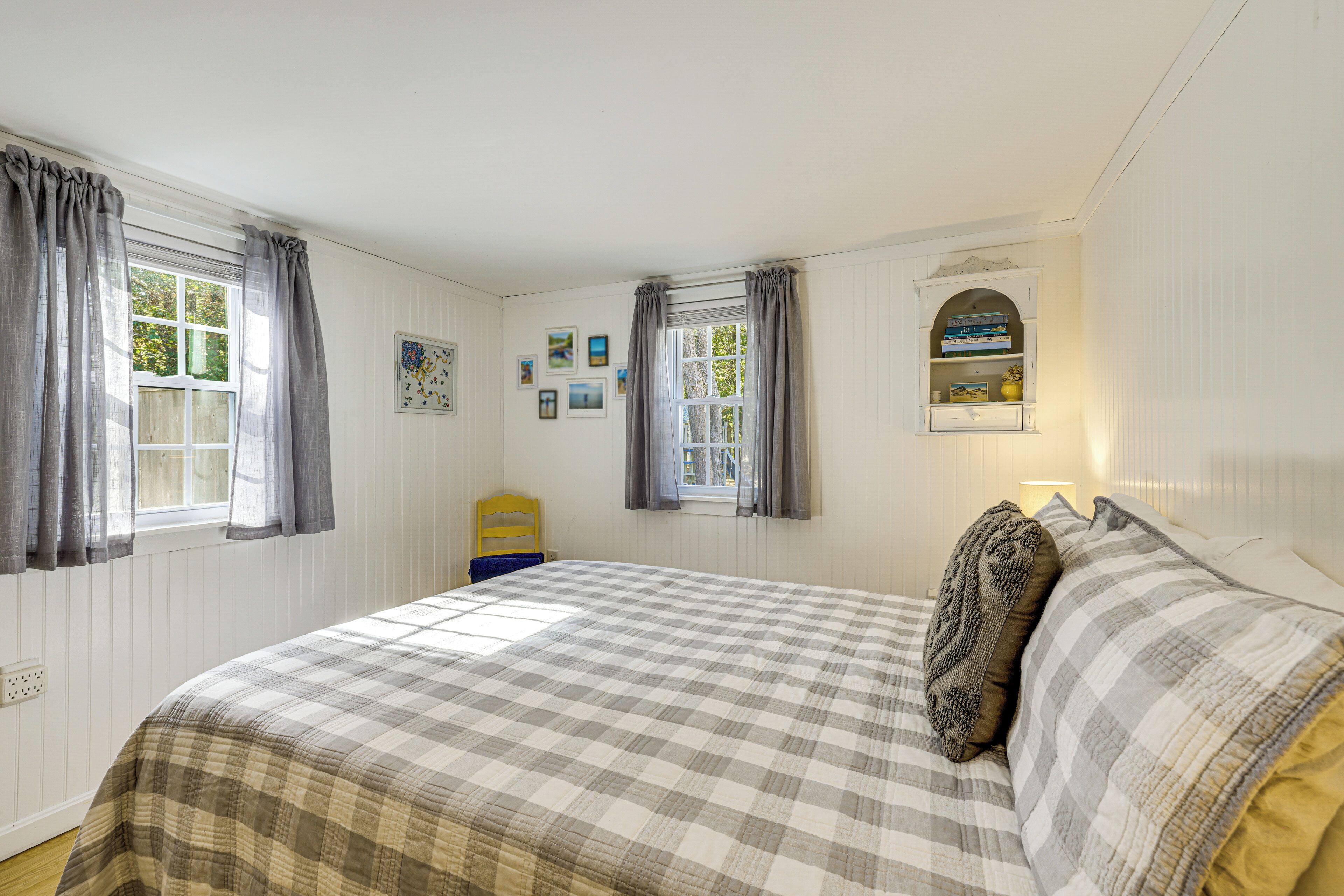 A bedroom with a large bed, two windows with curtains, a small shelf with books, and a wall-mounted picture frame.