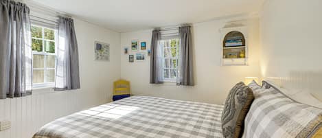 A bedroom with a large bed, two windows with curtains, a small shelf with books, and a wall-mounted picture frame.