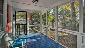 Eine überdachte Veranda mit blauem Boden, ein Korbstuhl mit Kissen und Blick auf einen Garten mit Bäumen und einer Holzterrasse.