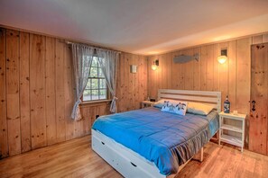 A wooden-paneled room with a bed, a nightstand, and a window with curtains. - Swim & Unwind! Follins Pond Cottage Near Beach (South Dennis)