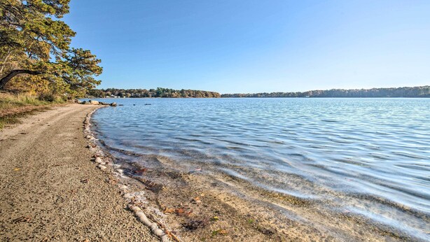 Ein Kiesweg führt zu einem ruhigen See mit klarem Wasser, umgeben von Bäumen und einer entfernten Uferlinie.
