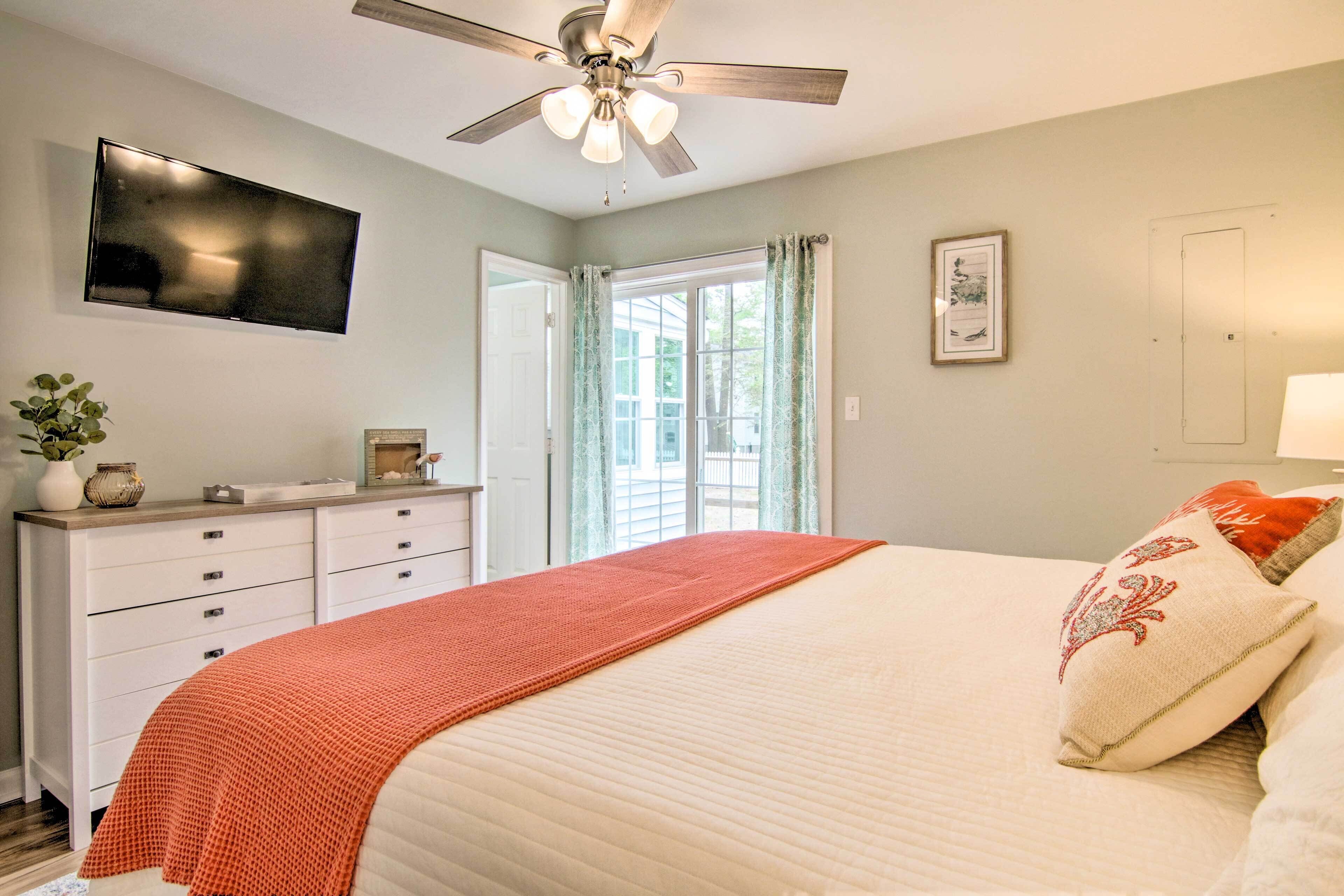Une chambre à coucher comprenant un lit, une commode, un téléviseur, un ventilateur de plafond et une fenêtre avec des rideaux.