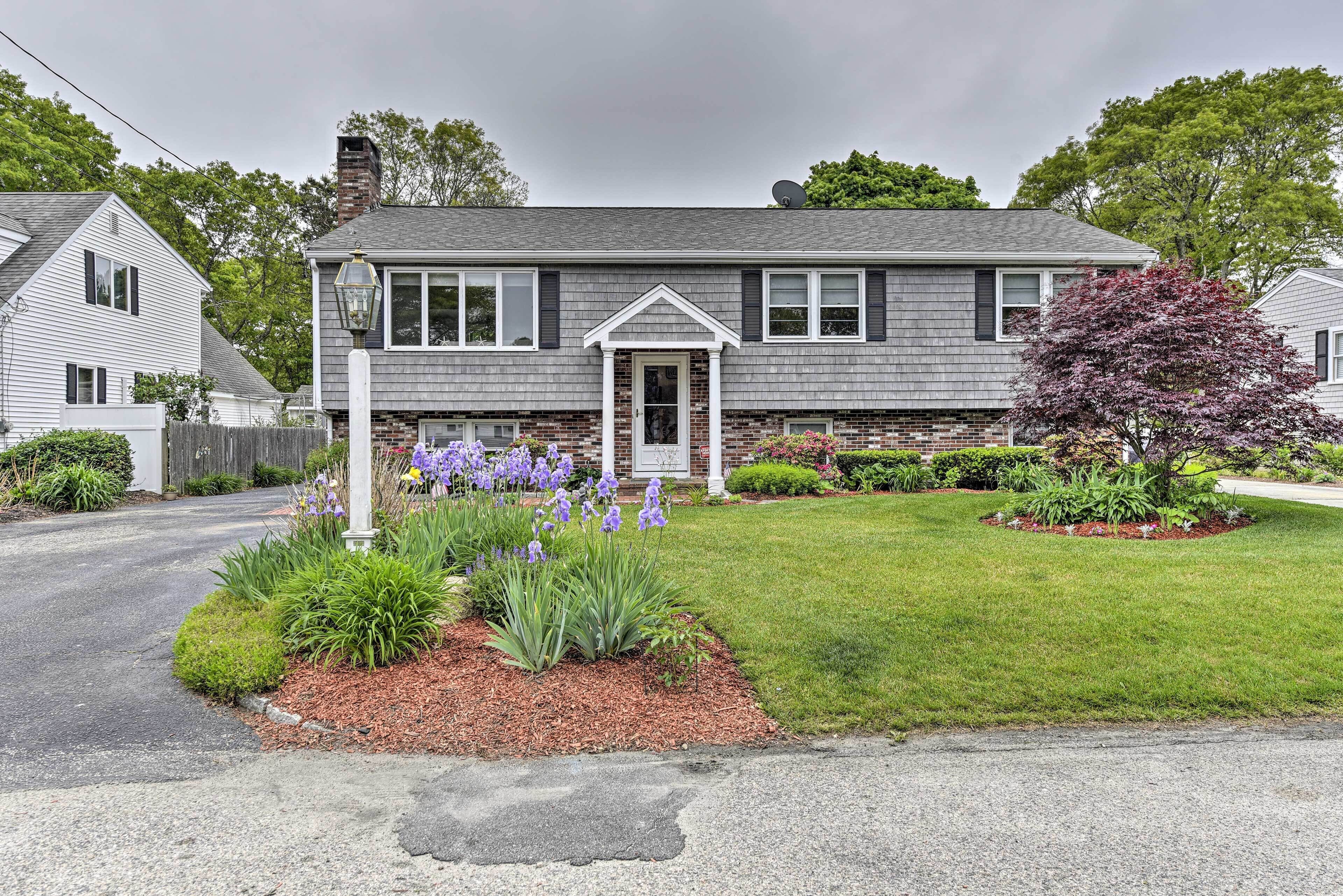 A two-story house with a front porch, a brick chimney, and a well-maintained lawn with purple flowers.