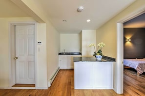 A compact kitchen with white cabinets, a granite countertop, and a small island. There is a bed with a striped bedspread in the adjacent room.