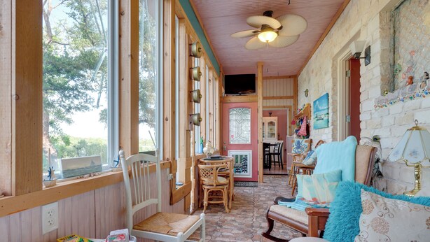 Una habitación acogedora con techo de madera, una zona de comedor con mesa y sillas, un sofá con cojines estampados y un ventanal con vistas a árboles.