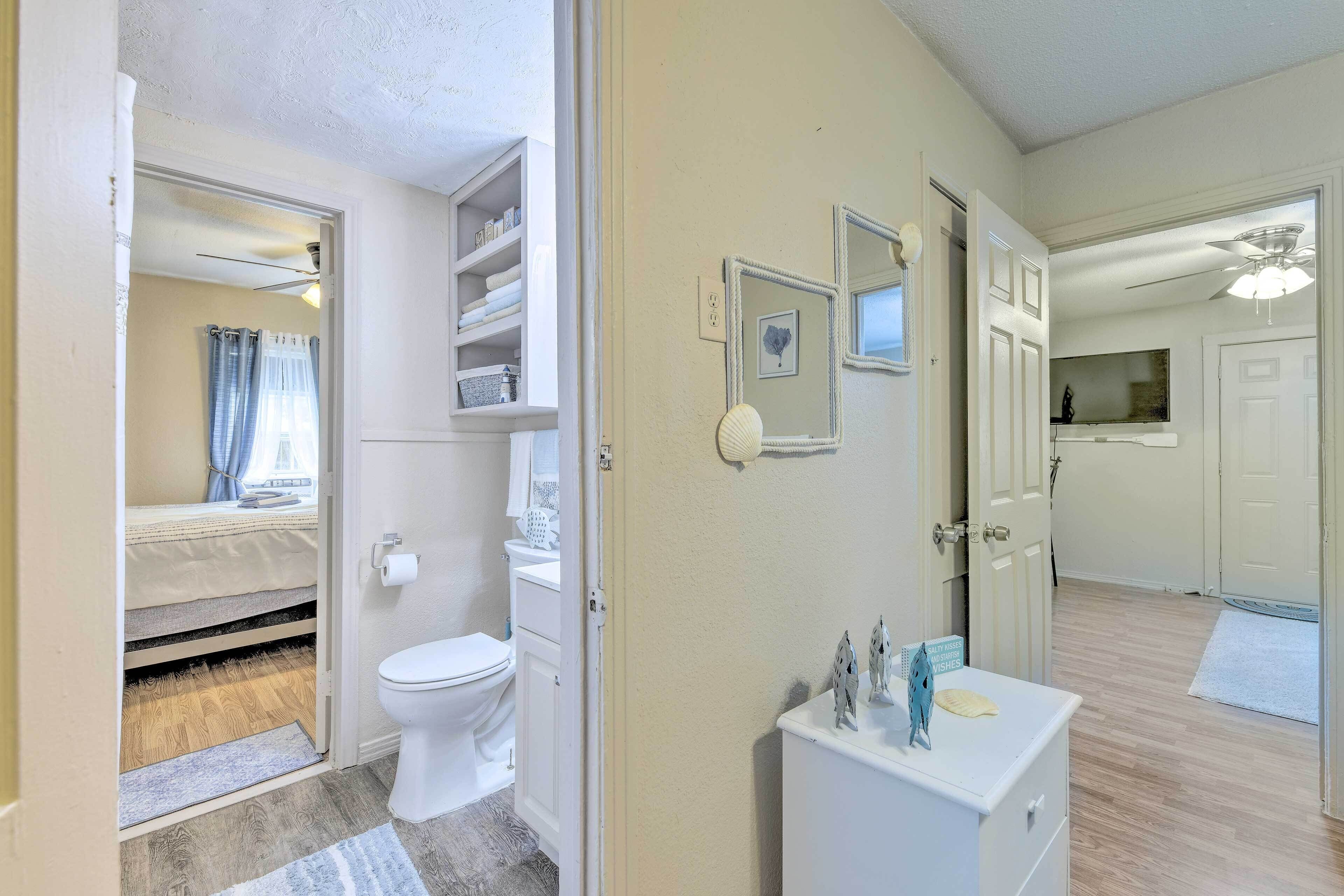 Habitación compacta y bien iluminada con inodoro, lavamanos y un mueble pequeño. Se ve una cama con cabecera a través de una puerta abierta, y un ventilador de techo en la habitación.