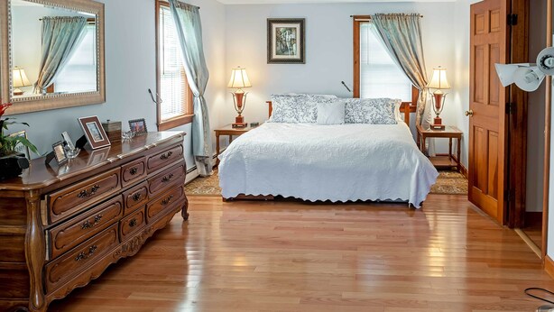 Quarto com cama grande, cómoda de madeira, duas mesas de cabeceira com luminárias, um quadro na parede e uma porta à direita.