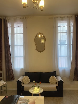 A living room with a sofa, a small round table with a vase and books, a wall-mounted mirror, and two windows with curtains. - ALBI-SUITE: Apartment in the heart of the city with free, private parking (Albi)