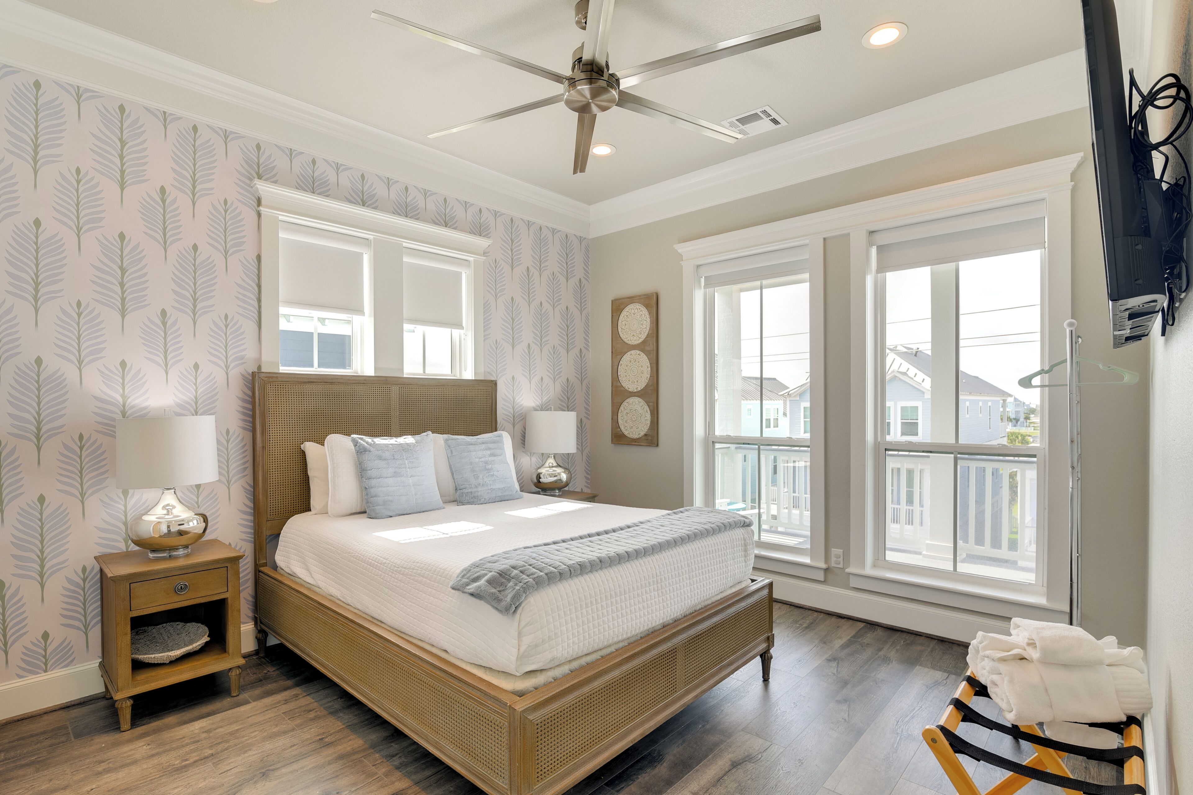 A bedroom with a large bed, two bedside tables, a ceiling fan, and a view of the outside through large windows.