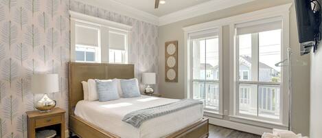 A bedroom with a large bed, two bedside tables, a ceiling fan, and a view of the outside through large windows.
