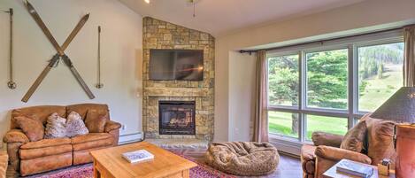 A cozy living room with a stone fireplace, leather furniture, and a large window offering a view of the outdoors.