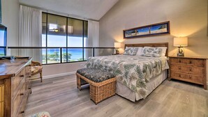 Un dormitorio con un ventanal grande, una cama con cubrecama de temática tropical, un aparador de madera y un banco de mimbre.