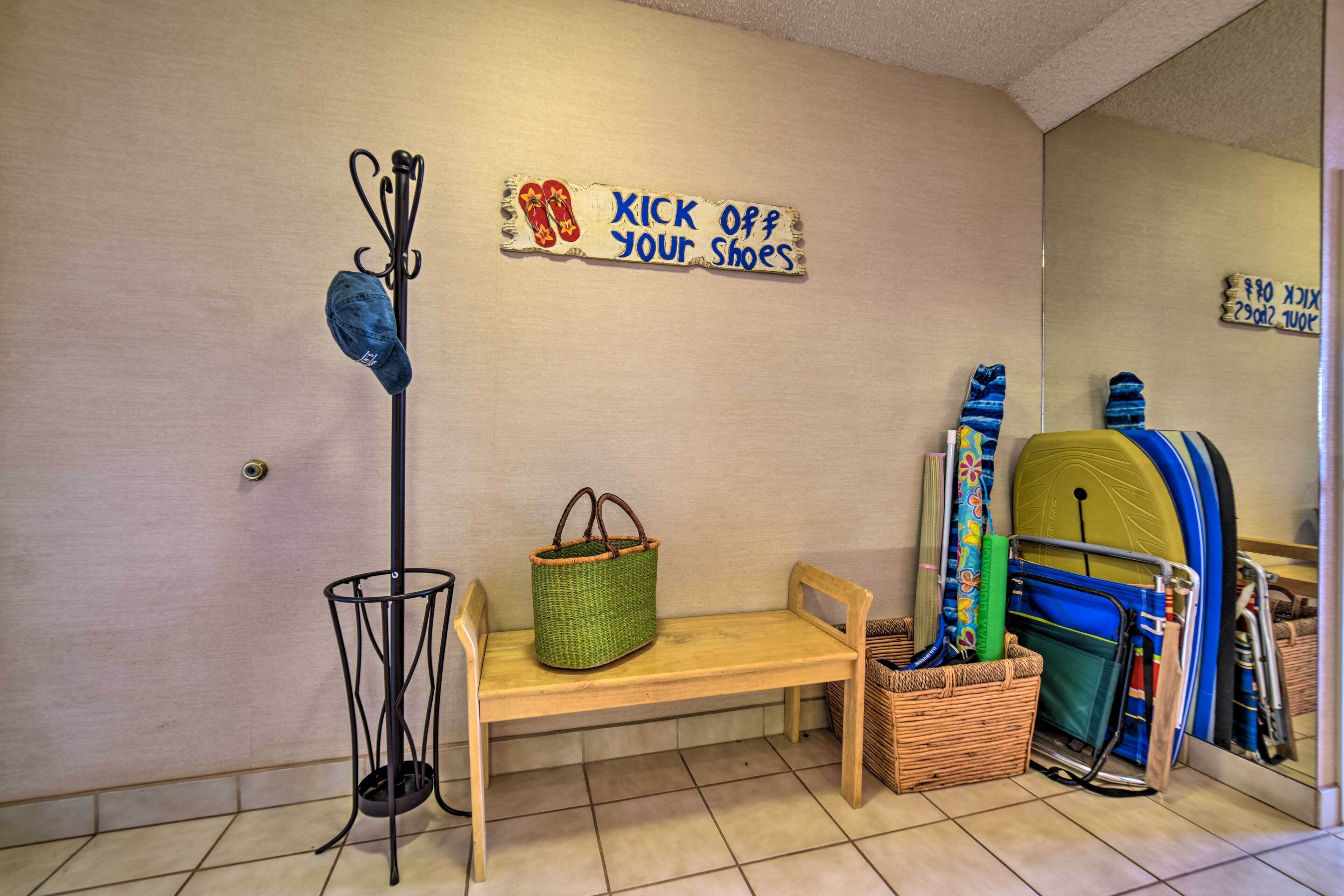 A room with a wooden bench, a coat rack with shoes, a green basket, and a wall-mounted sign that says "KICK OFF YOUR SHOES".