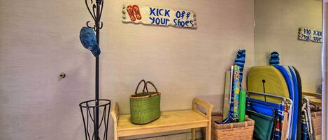 A room with a wooden bench, a coat rack with shoes, a green basket, and a wall-mounted sign that says "KICK OFF YOUR SHOES".