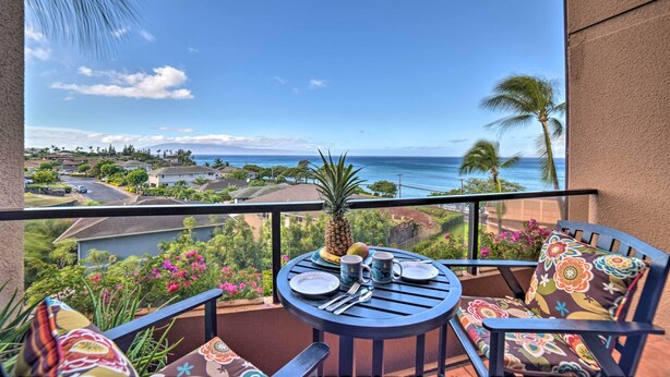 Un balcón con una mesa para dos, con vistas a la costa, casas y palmeras.