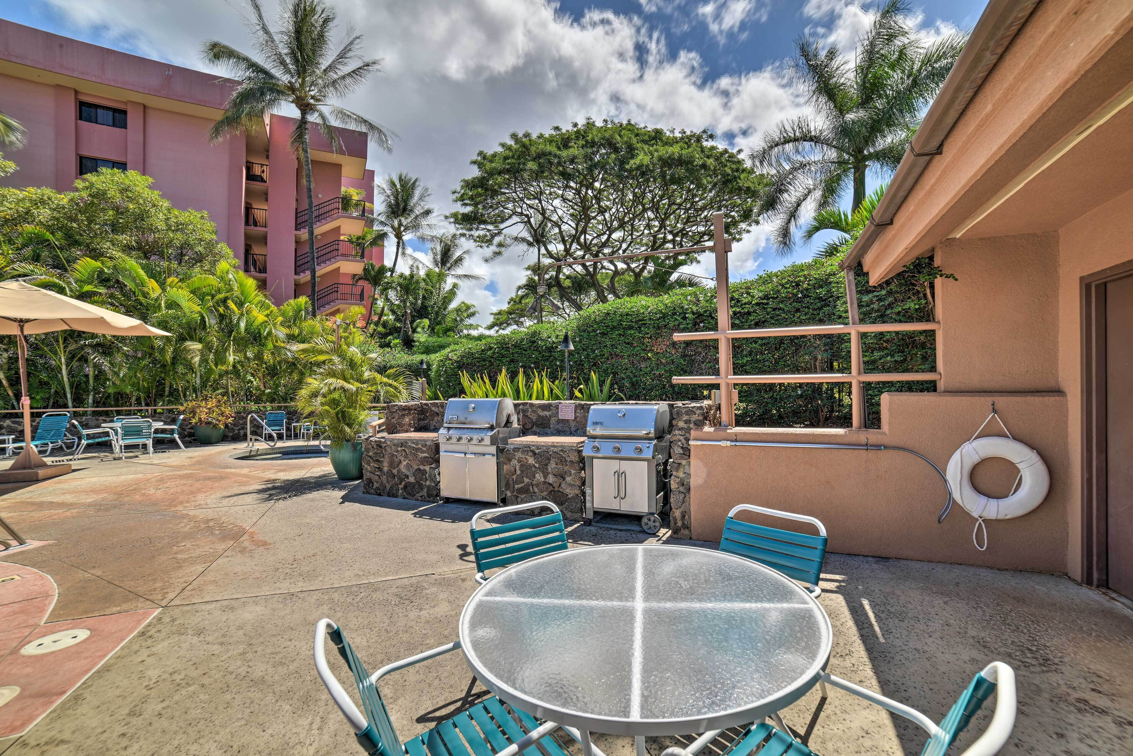 An outdoor patio with a round glass table and blue chairs, surrounded by lounge chairs and a barbecue grill.