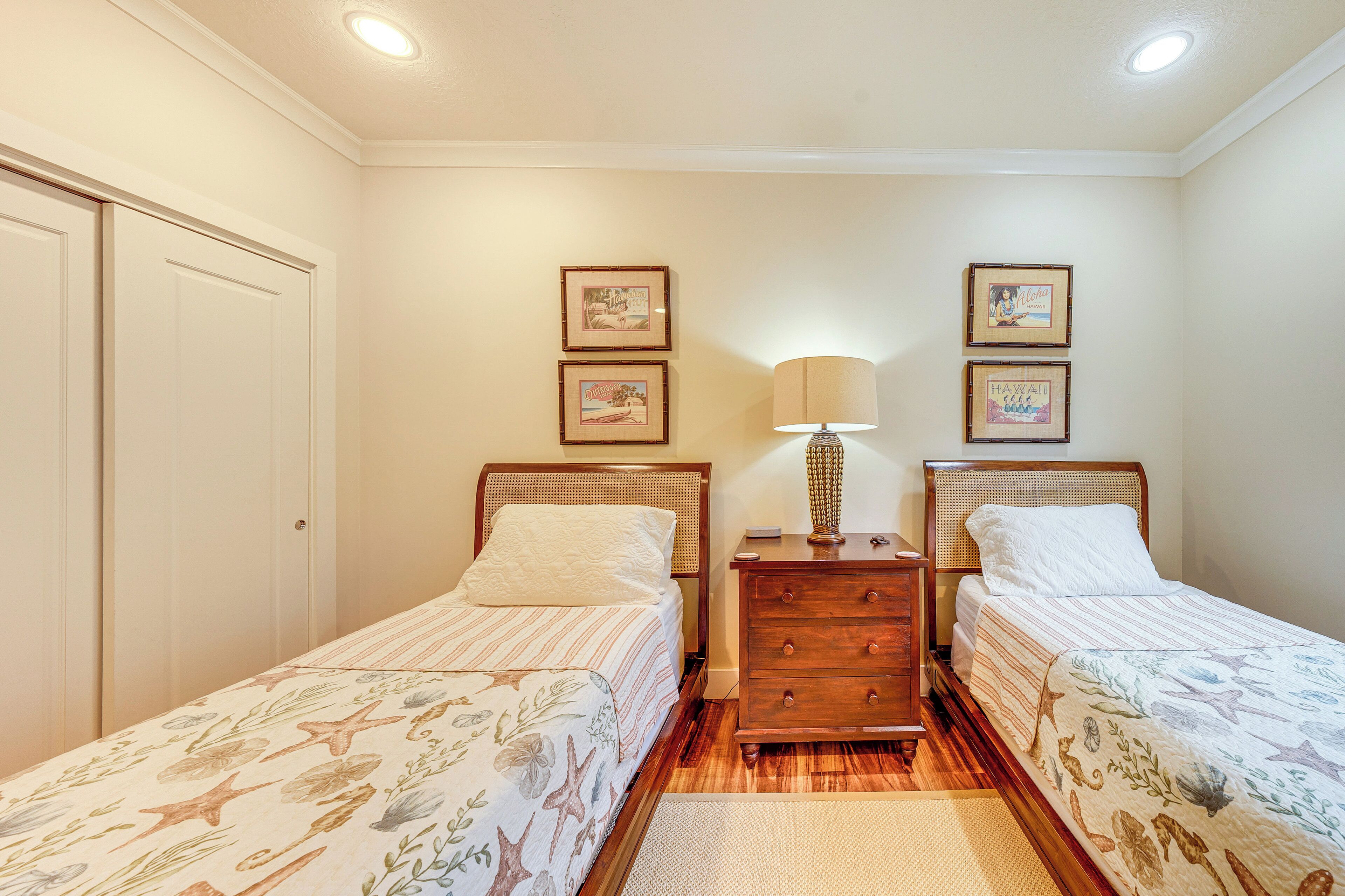 Une chambre avec deux lits simples, une commode en bois, une table de chevet avec une lampe et des tableaux encadrés au mur.