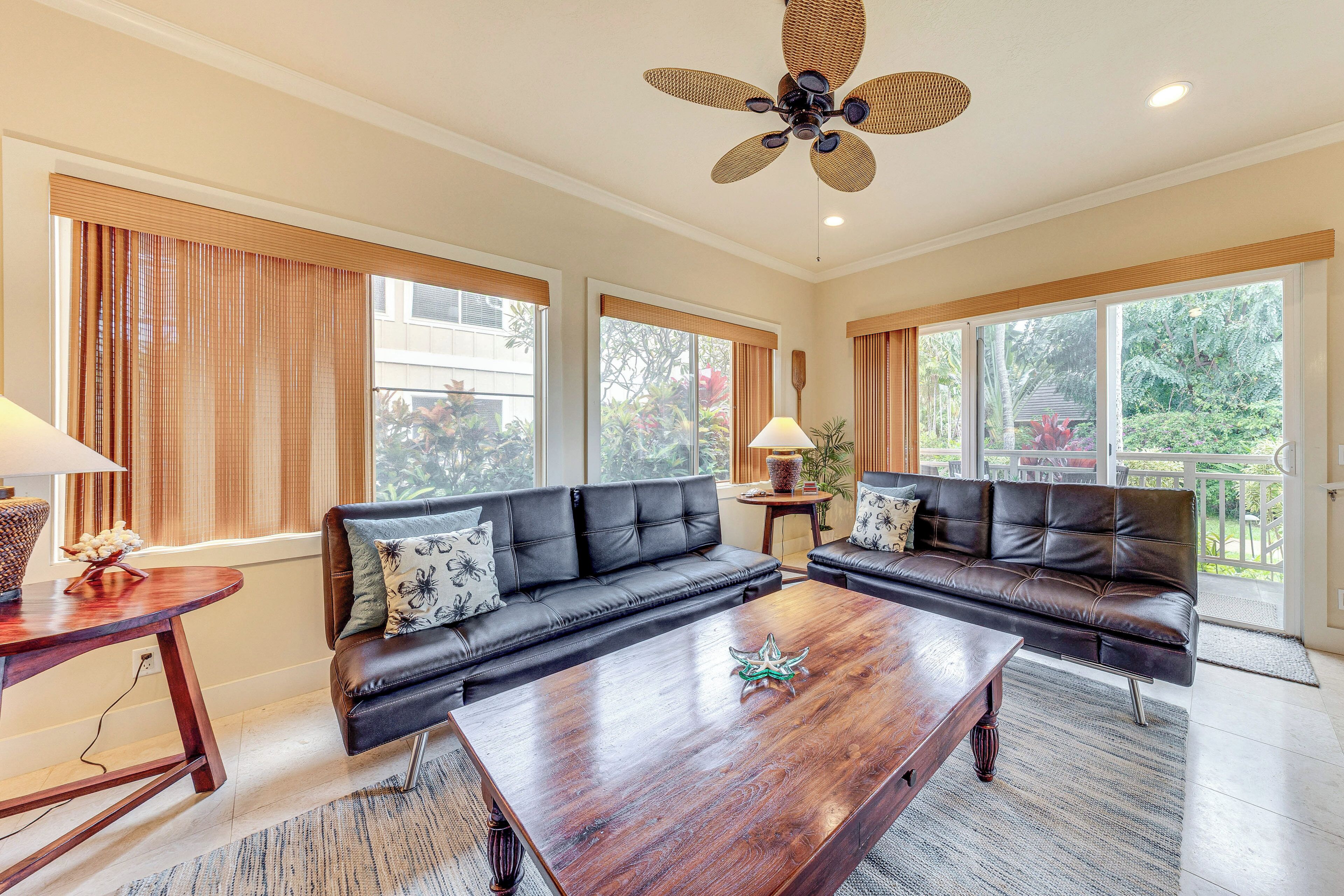 A spacious living room with two black leather sofas, a wooden coffee table, and a ceiling fan. Large windows provide natural light and views of greenery outside.