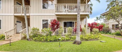 A two-story building with a balcony, a staircase, and a well-maintained lawn with tropical plants.