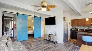 A modern living room with a sofa, a dining table, a kitchen area with wooden cabinets, a TV mounted on the wall, and a ceiling fan. - < 1 Mi to Beach + Town! Airy Kailua-kona Condo (Kailua-Kona)