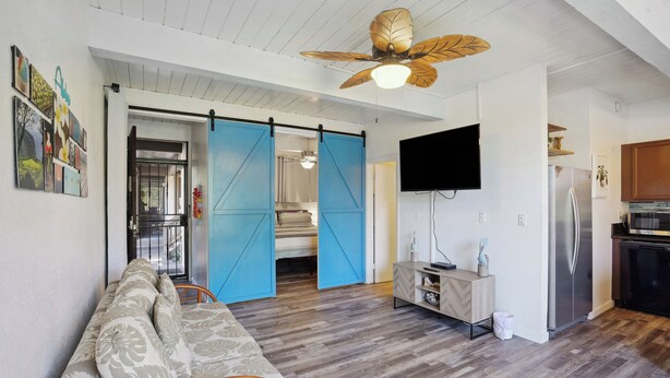 A modern living room with a sofa, a ceiling fan, a sliding door, a TV, and a kitchen area with a refrigerator. - < 1 Mi to Beach + Town! Airy Kailua-kona Condo (Kailua-Kona)