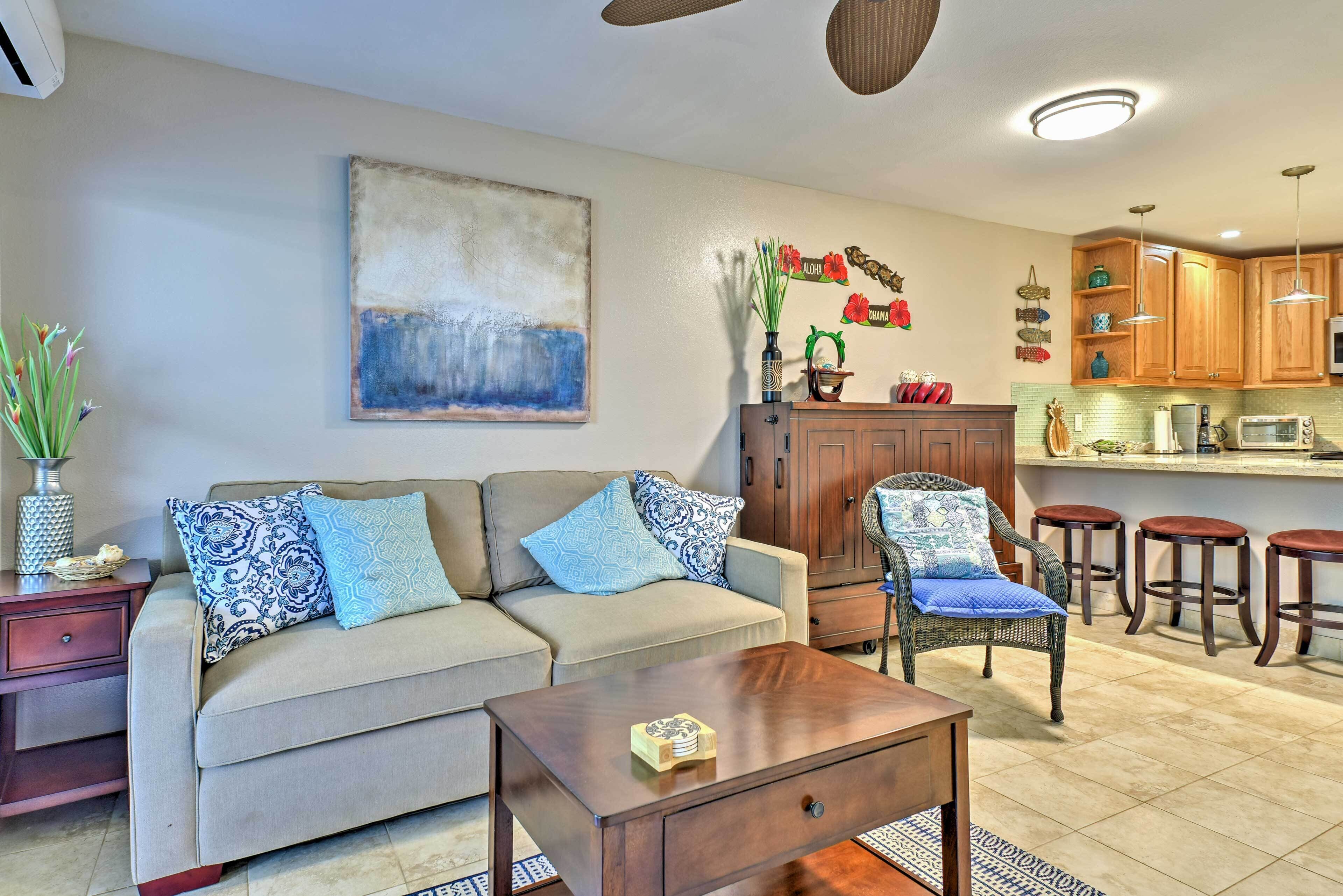 A living room with a sofa, coffee table, and a wicker chair. There is a kitchen area with wooden cabinets and a microwave.