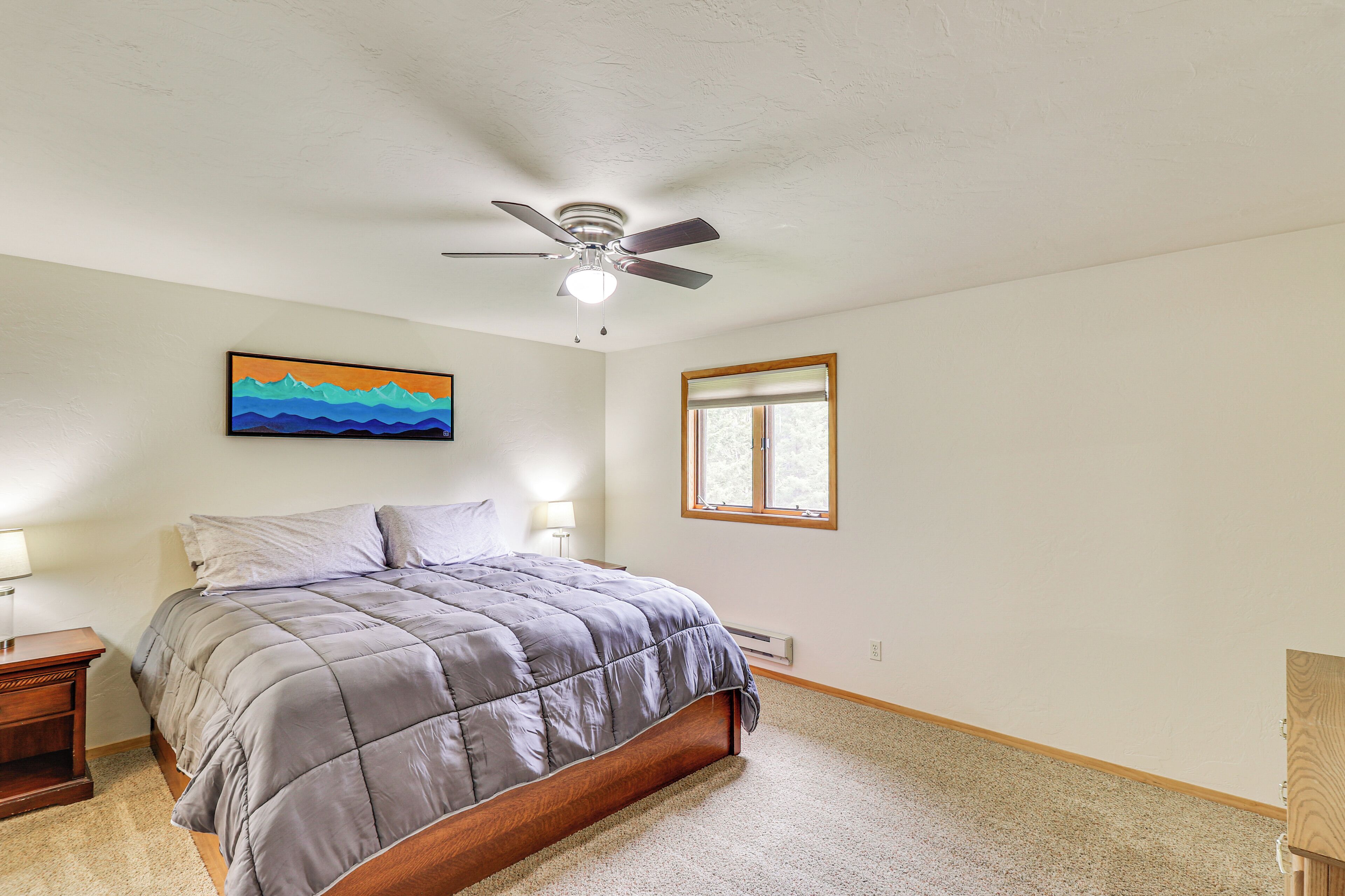 A bedroom with a bed, a ceiling fan, a window, and a painting on the wall.