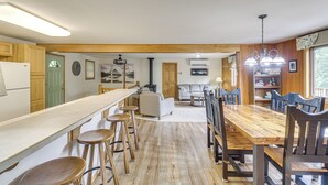 A spacious kitchen with wooden cabinets, a long counter, and bar stools. There is a dining area with a wooden table and chairs, and a living room with a sofa and a fireplace. - Scenic Forest Lodge Outside Glacier National Park! (West Glacier)