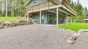 A two-story cabin with a balcony, stone wall, and a gravel driveway. - Scenic Forest Lodge Outside Glacier National Park! (West Glacier)