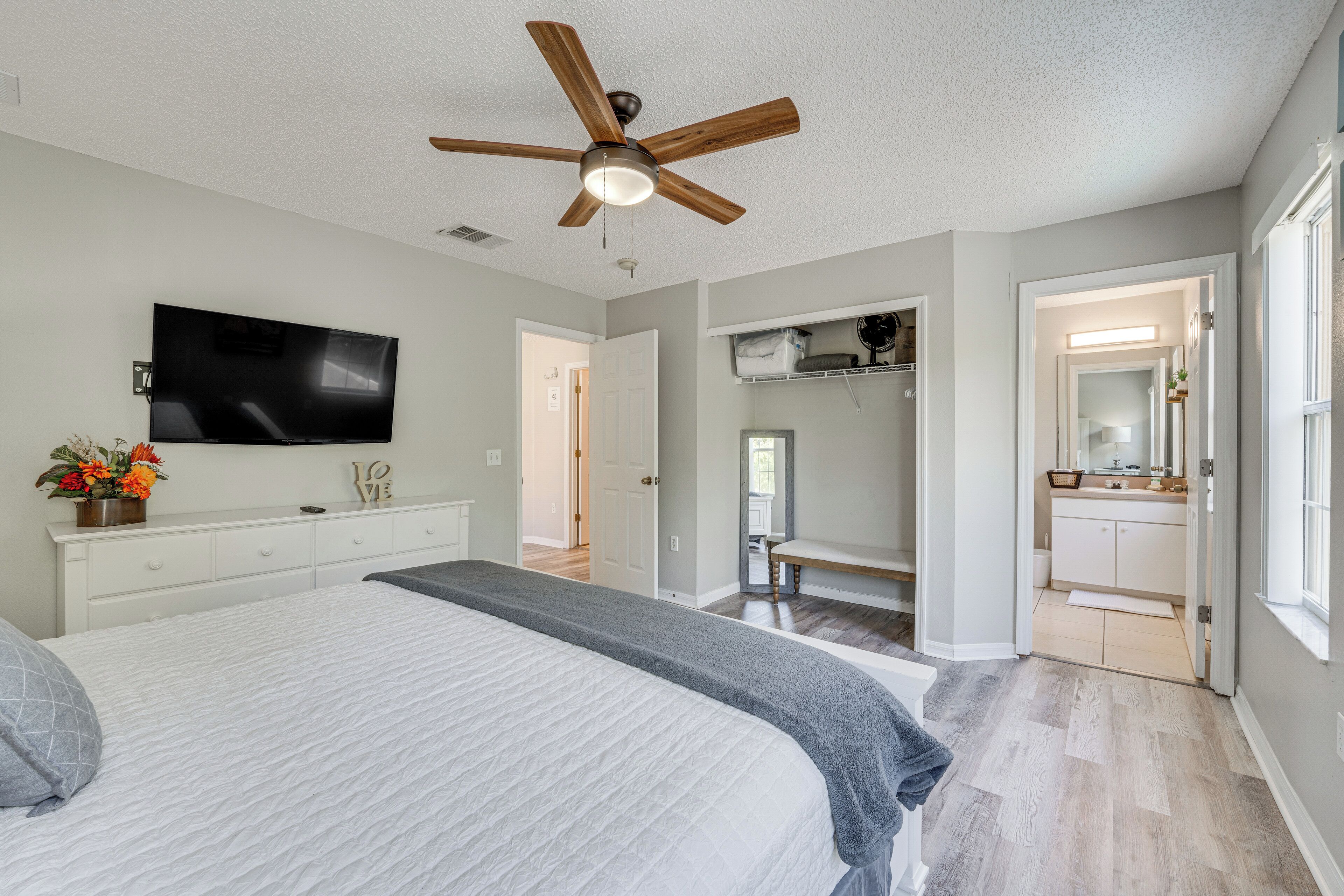 A bedroom with a large bed, a ceiling fan, a TV, and a bathroom visible through an open door.