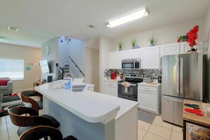 Une cuisine moderne avec des meubles blancs, un îlot central, des appareils en acier inoxydable et un coin repas comprenant un canapé gris et des chaises en bois.
