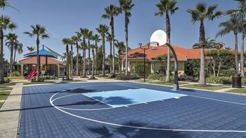 Una cancha de baloncesto con superficie azul, rodeada de palmeras y un edificio con tejado rojo.
