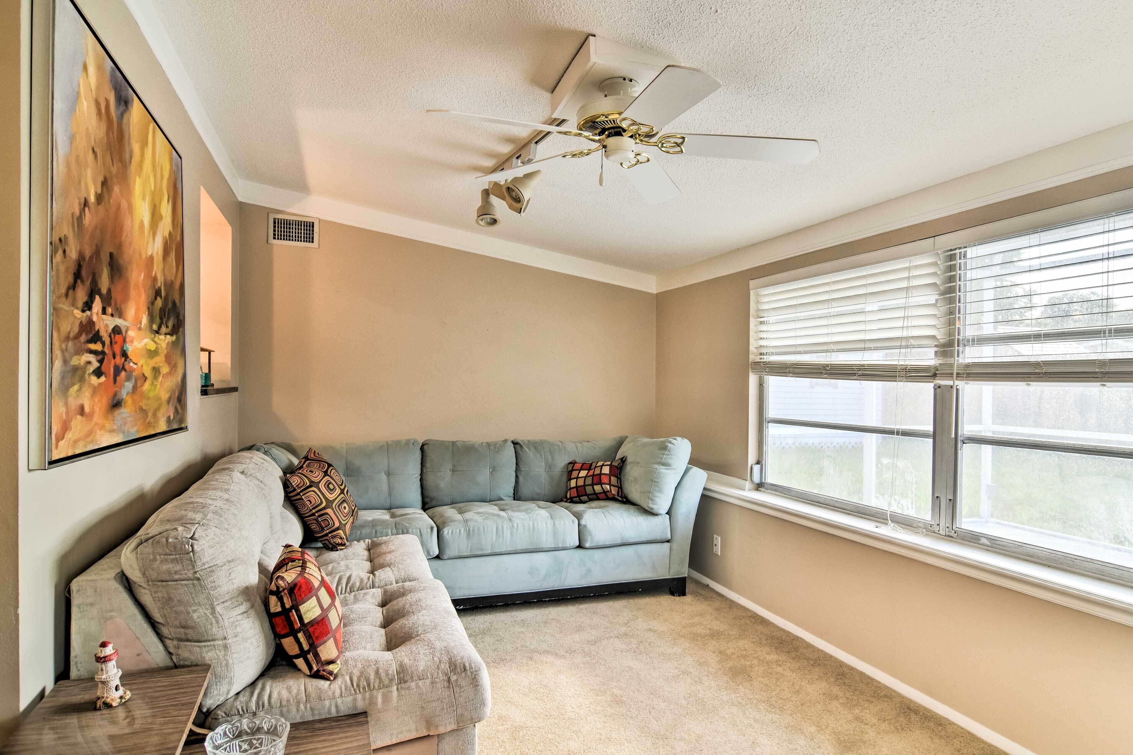 A living room with a gray sectional sofa, a ceiling fan, a large window with blinds, and a painting on the wall.