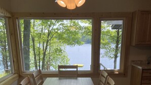 Zona pranzo con tavolo e sedie in legno, ampie finestre vista su lago e alberi, e una ventola a soffitto.