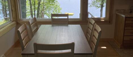 Ruokailutilassa on puinen pöytä ja tuolit, suuret ikkunat, joista avautuu näkymä järvelle ja puille, sekä kattotuuletin.